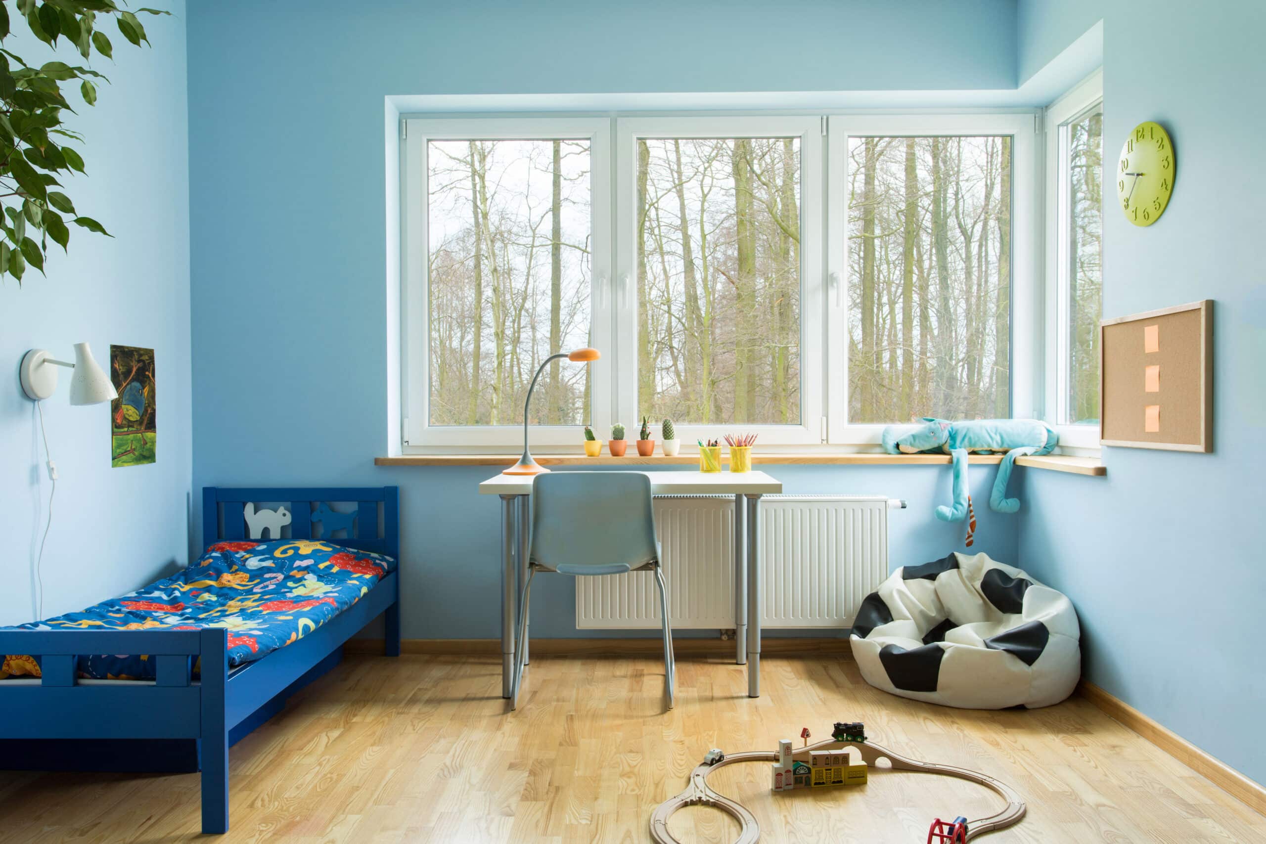 Light Blue Bedroom Interior Paint Light blue painted bedroom interior with white ceiling, blue bed frame, wooden desk, and large windows overlooking trees.